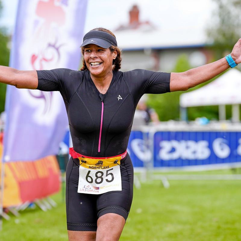 A person wearing a black triathlon suit and race bib number 685 smiles broadly with arms outstretched while crossing the finish line at an outdoor event. In the background, there are banners, spectators, and a grassy area.