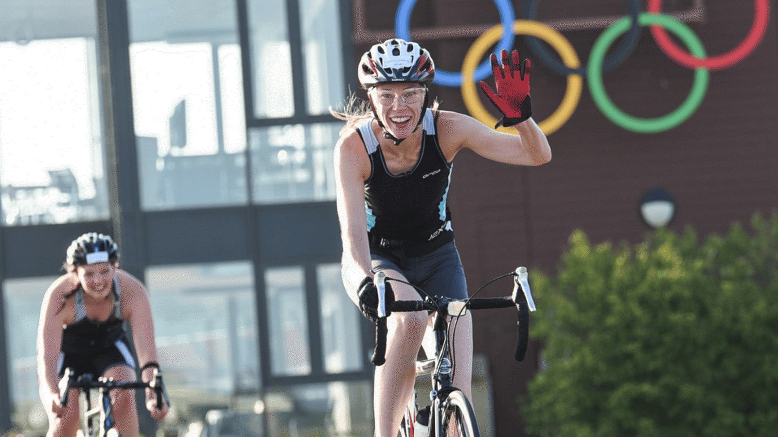 Two cyclists ride outdoors by a building with Olympic rings. The cyclist in front, wearing a helmet and red glove, smiles and waves. The other cyclist follows behind, also wearing a helmet.