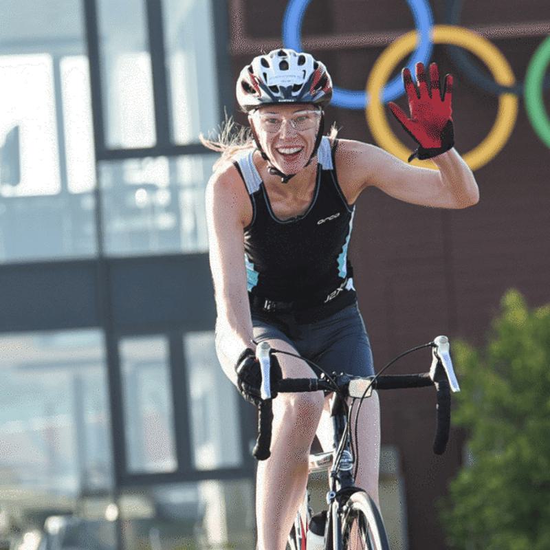 Two cyclists ride outdoors by a building with Olympic rings. The cyclist in front, wearing a helmet and red glove, smiles and waves. The other cyclist follows behind, also wearing a helmet.