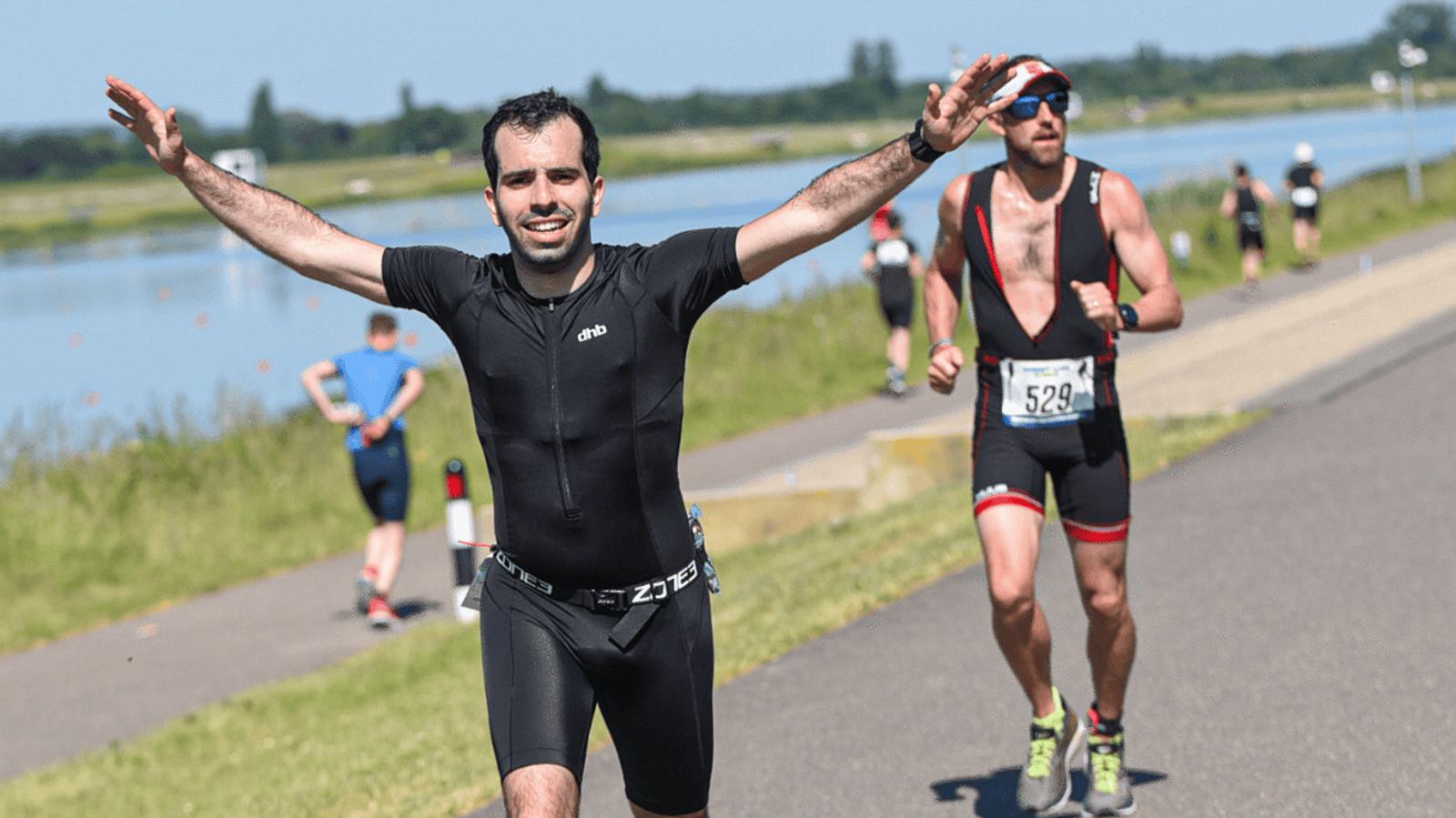 A man in a black athletic outfit raises his arms triumphantly while running on a paved path by a body of water. Other runners follow behind him. It's a sunny day, and the scene is outdoors.