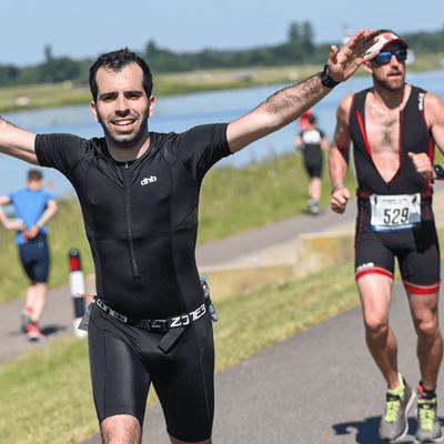 A man in a black athletic outfit raises his arms triumphantly while running on a paved path by a body of water. Other runners follow behind him. It's a sunny day, and the scene is outdoors.