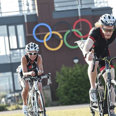 Two cyclists wearing helmets are racing on a sunny day. In the background, there's a building with the Olympic rings displayed. The cyclists are focused and riding on a clear path. Trees and greenery surround the area.