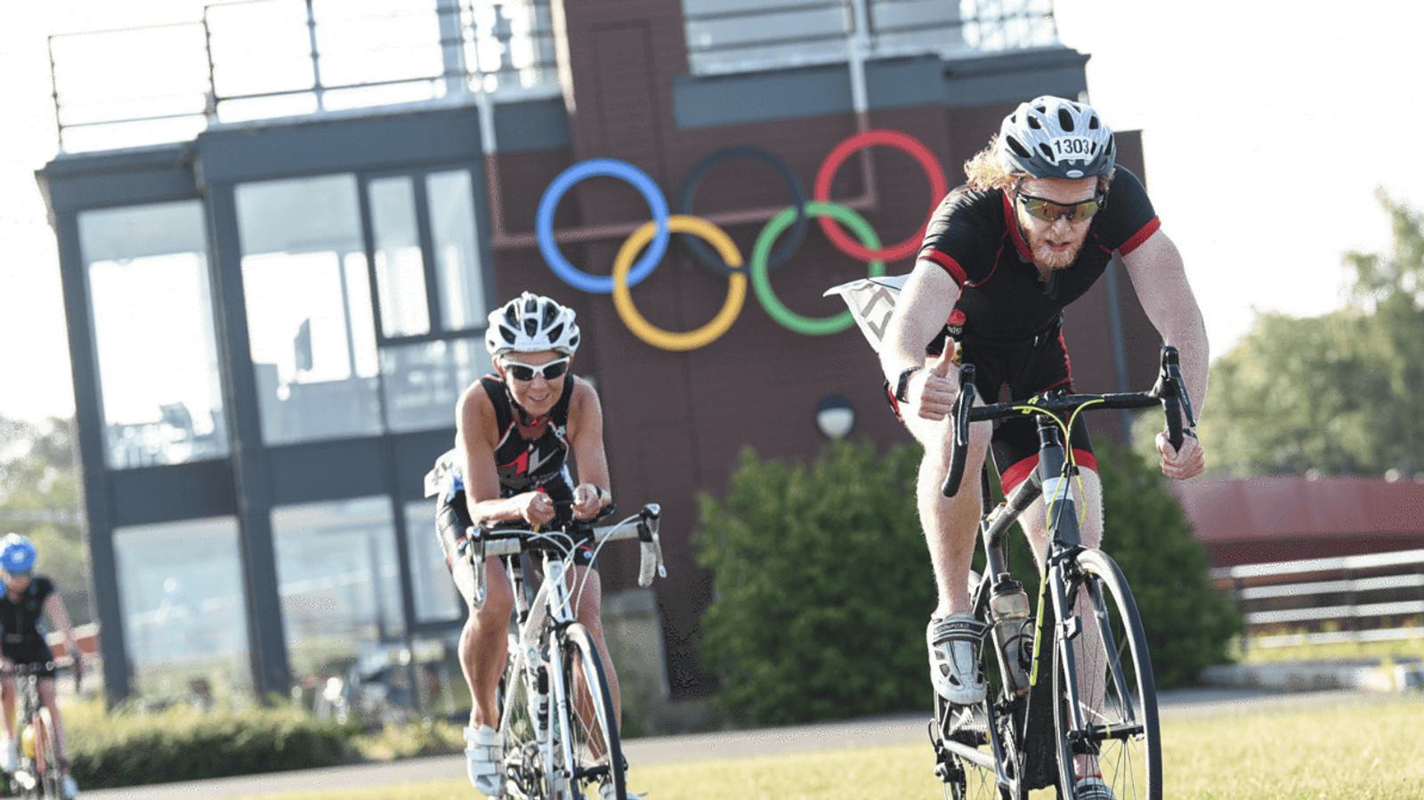 Two cyclists wearing helmets are racing on a sunny day. In the background, there's a building with the Olympic rings displayed. The cyclists are focused and riding on a clear path. Trees and greenery surround the area.