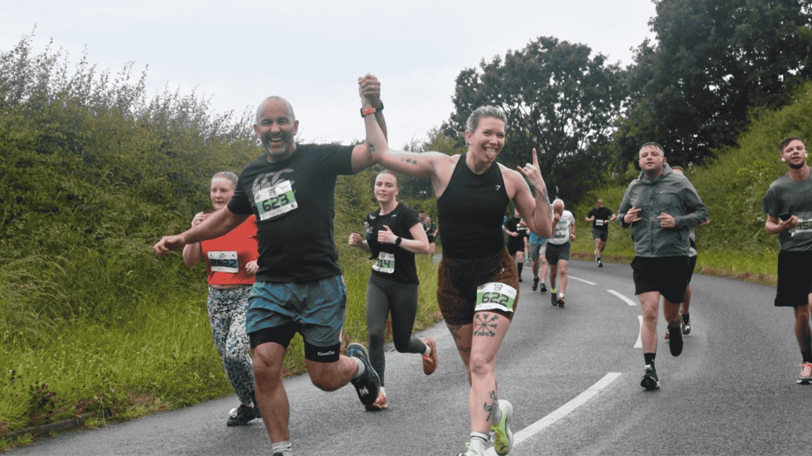 Runners participate in a race on a rural road. A man and woman hold hands, smiling and celebrating as they lead the group. Lush greenery and bushes line the road on both sides. Other runners follow behind, some focused and others cheerful.
