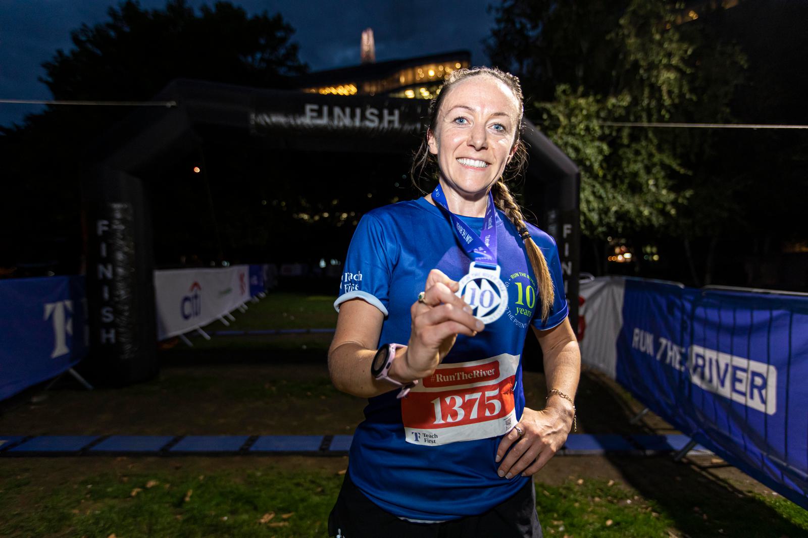 A runner in a blue jersey proudly displays her medal at the finish line of a race. She is wearing a race bib with the number 1375, and it is evening with a dark sky and lit trees in the background. She is smiling and appears to be content with her achievement.