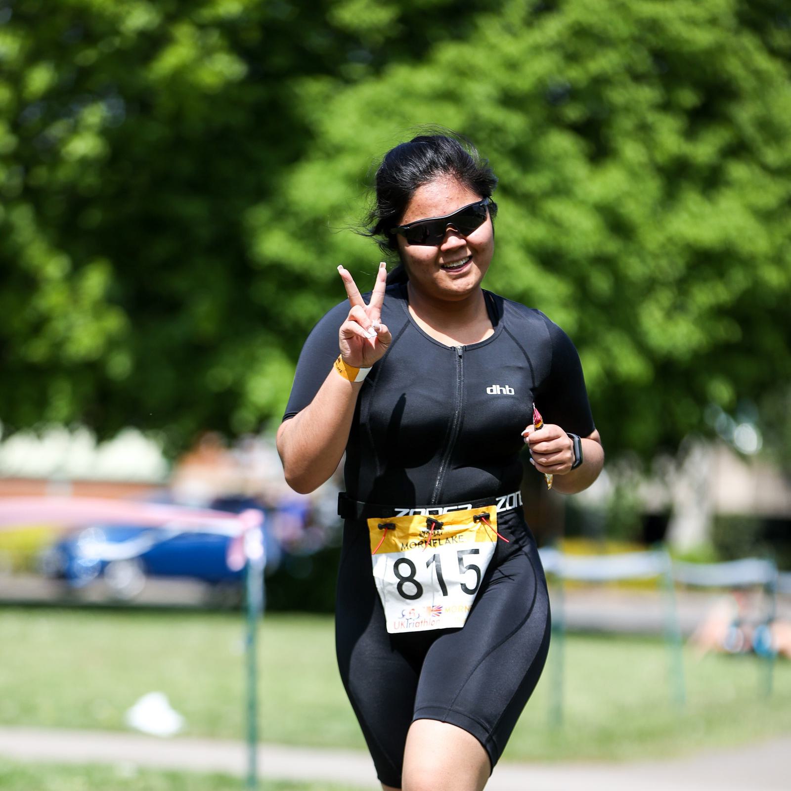 A person wearing sunglasses and a black athletic outfit is running outdoors, smiling, and giving a peace sign with their right hand. They are wearing a race bib with the number 815. The background features lush green trees and a sunny day.